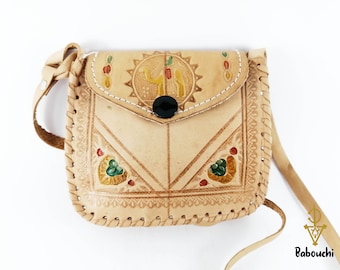 May include: A tan leather crossbody bag with a flap closure and a long strap. The bag features a camel design and decorative stitching. The bag is approximately 6 inches wide and 6 inches tall.