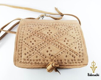 May include: A light tan leather crossbody bag with intricate geometric and floral embossed patterns. The bag features a long, adjustable strap and a small, rolled leather closure. The bag has the word "Babouchi" in the bottom right corner.