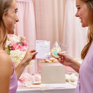 May include: Two people holding a pink and white invitation card and a cookie decorated with a blue design. The cookie is in a clear plastic bag. The invitation card says "Engaged".