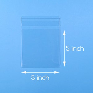 Peut inclure: Un sac en plastique transparent de forme rectangulaire, mesurant 12,7 cm sur 12,7 cm, sur un fond bleu clair. Le sac est transparent, permettant de voir le contenu. Les dimensions sont indiquées par des flèches et du texte blancs.