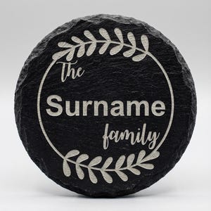 Puede incluir: Un posavasos redondo de pizarra negra con un diseño grabado en blanco. El diseño incluye las palabras "The Surname family" dentro de un borde circular con detalles de hojas. El posavasos tiene una superficie natural y texturizada.