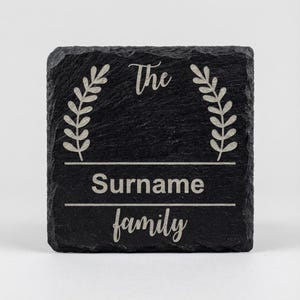 Puede incluir: Un posavasos cuadrado de pizarra negra con texto y diseño grabados. El texto dice "The Surname family" con un diseño de hojas decorativas a cada lado. Un regalo familiar personalizado.