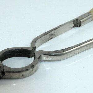 Könnte beinhalten: Ein Vintage-Metallwerkzeug mit einer Bürste an einem Ende und einer angelenkten, kreisförmigen Klemme am anderen. Das Werkzeug hat ein Paar Griffe zum Greifen und Ausüben von Druck. Die Bürste scheint aus Naturborsten zu bestehen.