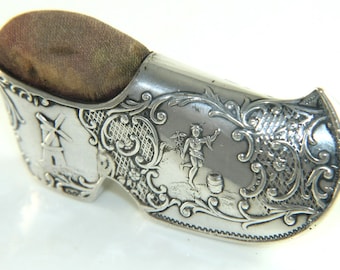 May include: Silver pincushion shaped like a shoe with an ornate design and a small windmill on one side and a cherub figure on the other.