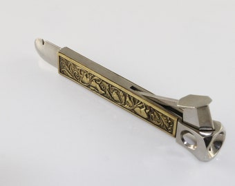 Peut inclure: Un coupe-cigare en argent et or avec un motif floral orné. Le coupe-cigare possède une lame pour couper les cigares et un poinçon pour faire des trous dans les cigares.