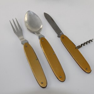 May include: A gold-handled pocket knife with a corkscrew, a spoon, and a fork. The knife has a silver blade and a gold handle.