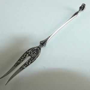 Pode incluir: Um garfo de prata com um design ornamentado e uma alça decorativa. O garfo tem aproximadamente 18 cm de comprimento.