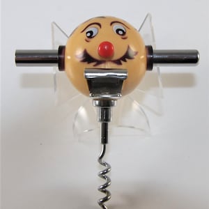 Op de afbeelding: Een novelty kurkentrekker met een lachend gezichtsontwerp. Het gezicht is beige met zwarte ogen, een rode neus en een zwarte snor. De kurkentrekker heeft een zilveren metalen worm en twee zilveren metalen handvatten.
