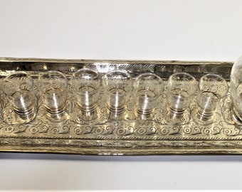 May include: A silver tray with an ornate design holds eight small clear glass shot glasses and a larger clear glass decanter. The tray has decorative handles on each end.