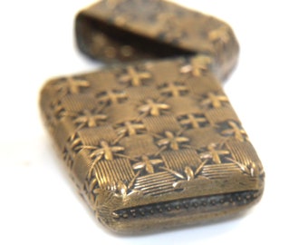 Könnte beinhalten: Eine goldfarbene Metall-Streichholzschachtel mit einem geprägten Blumenmuster auf der Oberfläche. Die Streichholzschachtel ist geschlossen und hat einen Klappdeckel.