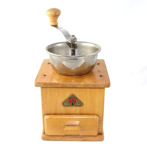 Könnte beinhalten: Eine antike Kaffeemühle aus Holz mit einem Metalltrichter und einem Kurbelgriff. Die Mühle hat eine Schublade zum Auffangen des Kaffeesatzes und ein Etikett mit der Aufschrift "De Ve" und einem Windmühlenlogo.