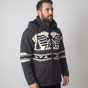 Puede incluir: Una sudadera de punto con cremallera en color gris carbón que presenta un gráfico de águila blanca en el pecho y los hombros. La prenda incluye un cuello grueso de canalé, mangas largas con rayas horizontales blancas y negras y dos bolsillos.