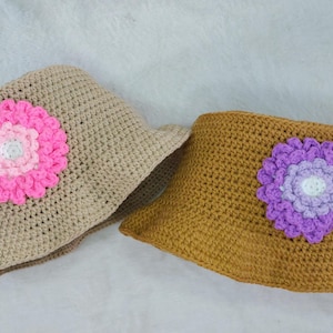 May include: Two crocheted bucket hats with flower embellishments. One hat is beige with a pink flower, the other is brown with a purple flower.