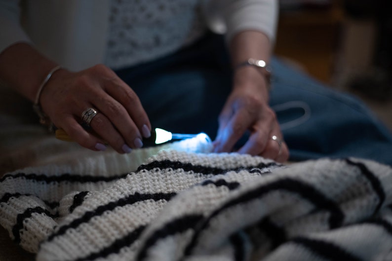 May include: A person is knitting a white and black striped crocheted blanket with a lighted needle.