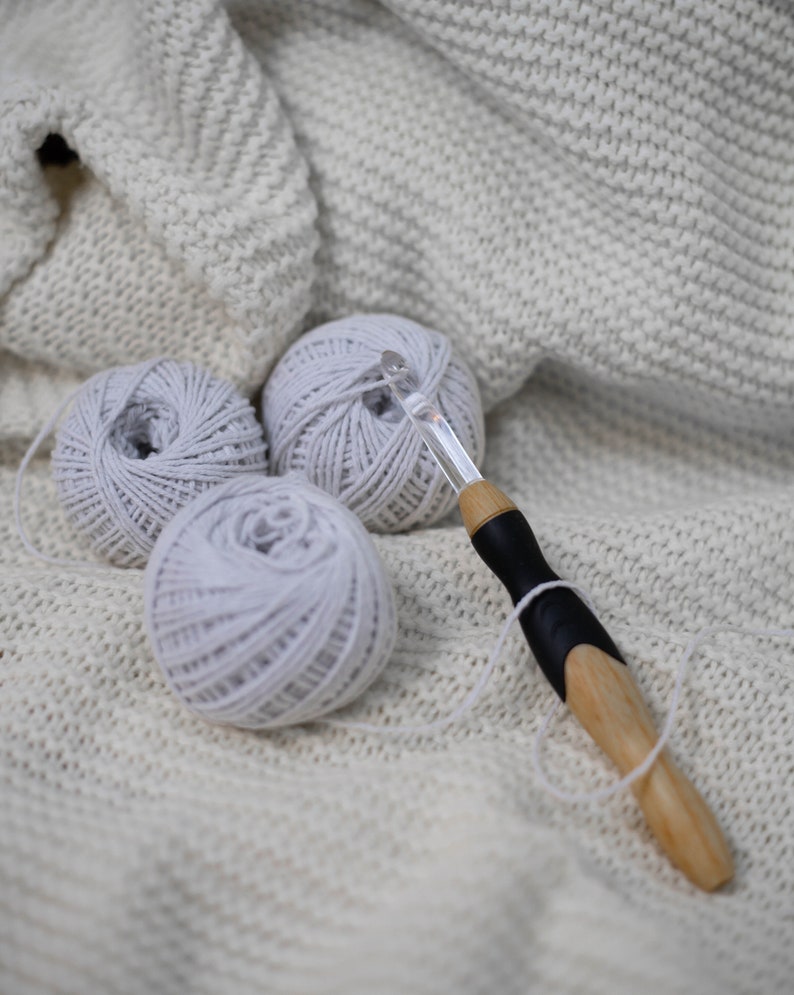 May include: Three balls of white yarn and a wooden crochet hook with a black handle on a white knitted blanket.