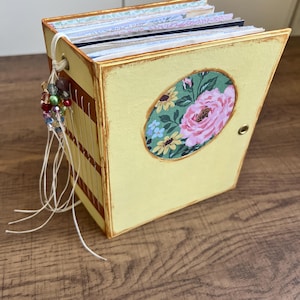 May include: A handmade yellow scrapbook album with a floral oval design on the cover. The album has a decorative beaded tassel and contains multiple patterned pages. The edges are painted with a brown trim.