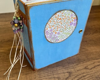 May include: A handmade, blue, wooden book with a gold trim and a floral oval design on the front. The book has a beaded charm and white string closure. The pages are visible, and the book is sitting on a wooden surface.