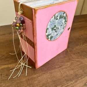 May include: A handmade pink journal with a gold-painted frame and a floral oval design on the cover. The journal is adorned with a beaded tassel and white string. The pages are visible, and the journal is sitting on a wooden surface.