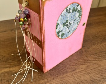 May include: A handmade pink journal with a gold-painted frame and a floral oval design on the cover. The journal is adorned with a beaded tassel and white string. The pages are visible, and the journal is sitting on a wooden surface.