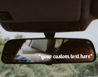 May include: A rearview mirror with a white sticker that says "*your custom text here*" on it. The mirror is reflecting a view of a road with trees on either side.