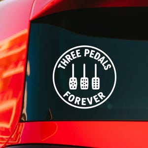 May include: A white circular vinyl decal applied to a car window. The design features a graphic of three car pedals in the center, surrounded by the text "THREE PEDALS" at the top and "FOREVER" at the bottom, all in a bold, sans-serif font.