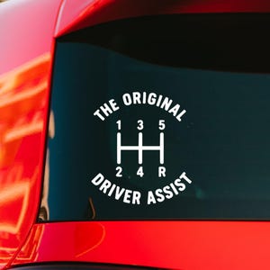 May include: A white vinyl decal applied to a car window featuring a manual transmission gear shift pattern with numbers 1, 3, 5, 2, 4, and R. The text THE ORIGINAL DRIVER ASSIST is arched around the gear diagram in a bold, sans-serif font.