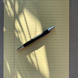 Puede incluir: Un bloc de notas amarillo con líneas y un bolígrafo negro encima. El bloc de notas está sobre una superficie gris texturizada.