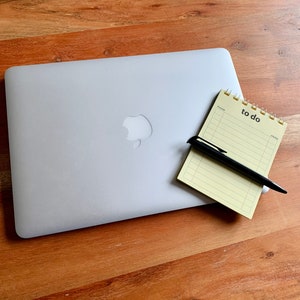 May include: A silver laptop computer with a white apple logo on a wooden table. A yellow notepad with the words "to do" written on it and a black pen are next to the laptop.