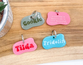 May include: Four irregular, blob-shaped pet ID tags with metal rings. One sage green tag reads Cookie, one pink tag has address text 012345 6789 10 Musterstraße 1A 12345 Musterstadt TassoNR 12345 678, one pink tag reads Tiida, and one blue tag reads Fridolin.