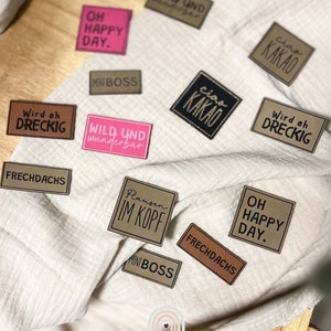 Könnte beinhalten: Eine Sammlung von rechteckigen und quadratischen Etiketten in verschiedenen Farben wie Pink, Braun, Schwarz und Beige. Die Etiketten tragen aufgedruckte Texte wie OH HAPPY DAY, WILD UND wunderbar, MINI BOSS, ciao KAKAO, Wird eh DRECKIG, FRECHDACHS und Flausen IM KOPF.