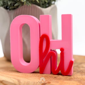 Könnte beinhalten: Ein freistehendes Dekorationsobjekt bestehend aus den großen rosa Blockbuchstaben O und H, wobei das Wort hi in roter Schreibschrift über das H gelegt ist. Die Buchstaben haben eine glatte, matte Oberfläche und sind zum Aufstellen gedacht.