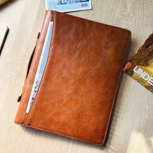 May include: A brown leather portfolio with a handle and a flap closure. The portfolio is open, revealing a white paper inside. The leather has a textured appearance. A magazine is partially visible in the background.