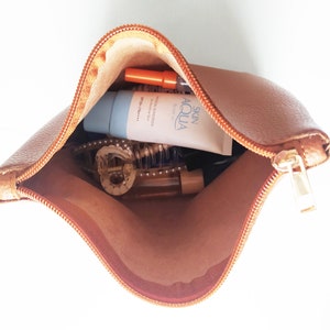 Puede incluir: Una bolsa de cuero marr&oacute;n con cremallera y un tirador de cremallera dorado. La bolsa est&aacute; abierta y contiene un tubo de protector solar azul y blanco, una botella de pl&aacute;stico transparente, una pinza para el pelo dorada y un resaltador rosa y naranja.