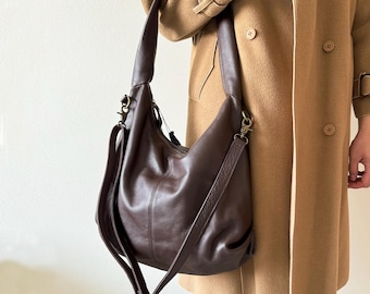 May include: A dark brown leather shoulder bag with a long adjustable strap and a shorter handle. The bag has a crescent shape and a zippered top. The bag is shown against a tan coat.