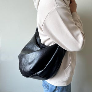 May include: A large, black, slouchy shoulder bag made of a textured material. The bag is worn over the shoulder, and the person in the image is wearing a cream-colored hoodie and blue jeans.