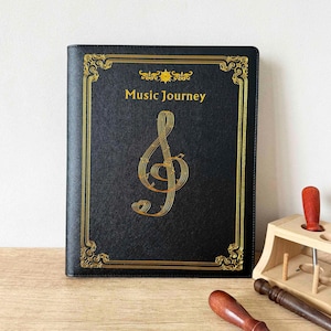 Könnte beinhalten: Schwarzes Notizbuch mit goldener Umrandung und einem goldenen Notenschlüssel-Design. Die Worte "Music Journey" sind in Gold gedruckt. Das Notizbuch liegt auf einer Holzoberfläche.
