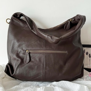 May include: A dark brown leather hobo bag with a soft, pebbled texture. The bag features a single wide shoulder strap, a top zipper closure with a leather pull, and a horizontal exterior zippered pocket on the front with a matching leather pull.