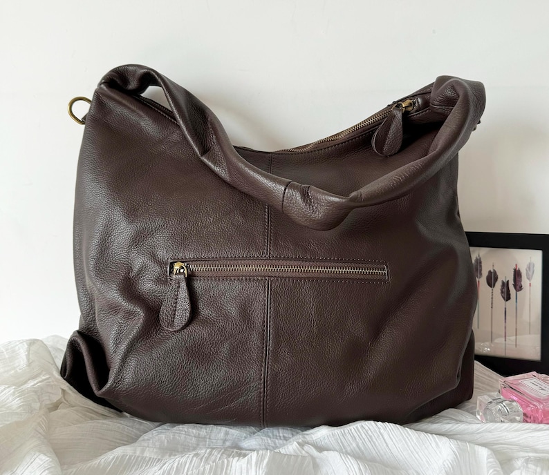 May include: A dark brown leather hobo bag with a soft, pebbled texture. The bag features a single wide shoulder strap, a top zipper closure with a leather pull, and a horizontal exterior zippered pocket on the front with a matching leather pull.