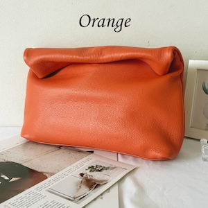 May include: A bright orange leather clutch bag with a soft, folded top closure and a pebbled texture. The word Orange is printed in black text above the bag. The design features a rectangular shape with rounded corners and a smooth, flexible finish.