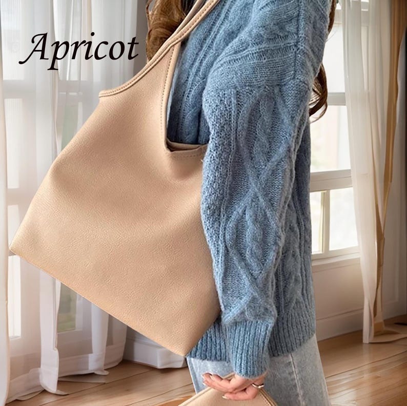 Puede incluir: Un bolso tote grande de color beige claro con un dise&ntilde;o holgado. El bolso est&aacute; hecho de un material suave y texturizado y tiene una parte superior ancha y abierta. El bolso lo lleva una persona que lleva un jersey de punto de cable azul claro y unos vaqueros azules.