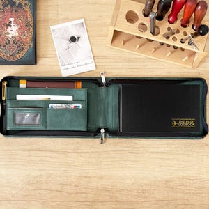 May include: A dark green zipped leather organiser case featuring multiple interior pockets for cards and documents, a gold pen, and a black notebook with the gold text THE PILOT LOGBOOK and an aeroplane icon printed on the front cover.