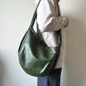 May include: A large, olive green shoulder bag with a curved, crescent shape. The bag has a long strap and a zipper closure. The leather-like material has a slightly distressed appearance. The bag is worn over the shoulder.