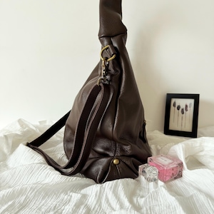 May include: A dark brown hobo bag with a pebbled texture, featuring a long adjustable shoulder strap attached with antique brass-toned metal hardware. The bag has a slouchy, unstructured silhouette with a single visible metal stud at the base.