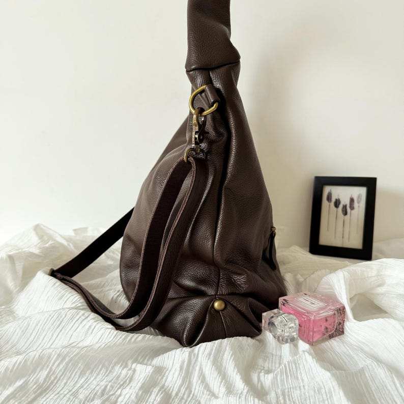 May include: A dark brown hobo bag with a pebbled texture, featuring a long adjustable shoulder strap attached with antique brass-toned metal hardware. The bag has a slouchy, unstructured silhouette with a single visible metal stud at the base.