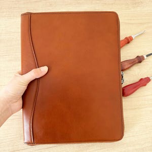 May include: A brown leather portfolio with a zipper closure. The portfolio is held by a hand and has a curved design on the left side. Three tools with wooden handles are visible.