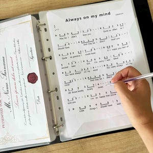 May include: A black binder with sheet protectors holding sheet music. The title "Always on my mind" is visible. A hand holding a pen is writing on the sheet music. A certificate is visible on the left.