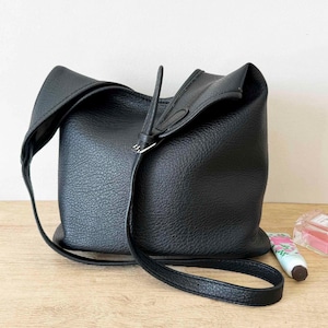 Puede incluir: Un bolso de mano de cuero negro con una superficie texturizada. El bolso tiene una correa larga y delgada y un diseño plegado único en la parte superior. El bolso está sobre una superficie clara.