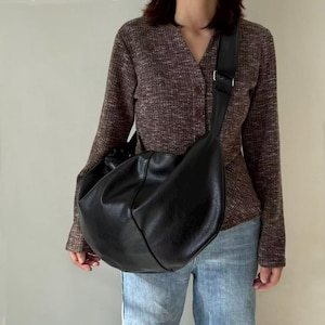 May include: A large, black shoulder bag with a wide strap. The bag is made of a soft, textured material and has a rounded shape. The strap is adjustable with a silver buckle. The bag is worn over the shoulder.