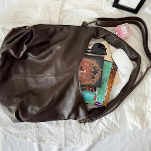 May include: A dark brown leather hobo bag with a long shoulder strap, containing a white computer mouse, black sunglasses, a small pink perfume bottle, and several books with colorful covers including one with a butterfly illustration.