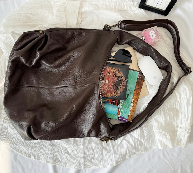 May include: A dark brown leather hobo bag with a long shoulder strap, containing a white computer mouse, black sunglasses, a small pink perfume bottle, and several books with colorful covers including one with a butterfly illustration.